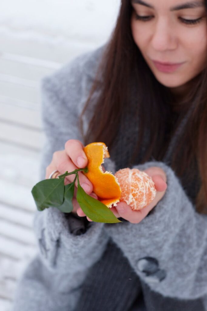 Woman in winter attire peeling a fresh tangerine outdoors, conveying freshness and seasonal joy.